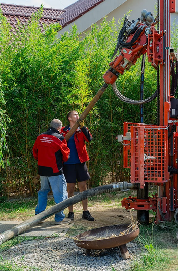 Dva muži v červených bundách REVER obsluhují malou vrtnou soupravu v osazené zelené zahradě, při vrtání studny.