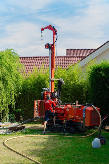 Červená vrtná souprava na pásech, pro vrtání studny a geotermálních vrtů, stojí na travnatém osazeném pozemku u domu a obsluhuje ji muž v budně REVER.