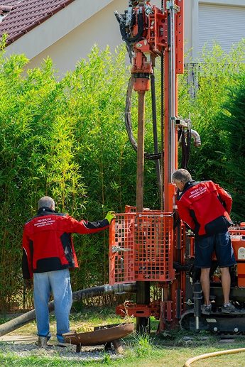 Dva muži v červených bundách REVERZ.CZ obsluhují červenou vrtací soupravu na zeleném trávníku. Stroj vrtá studnu nebo geotermální vrt. V pozadí je hustý zelený porost.