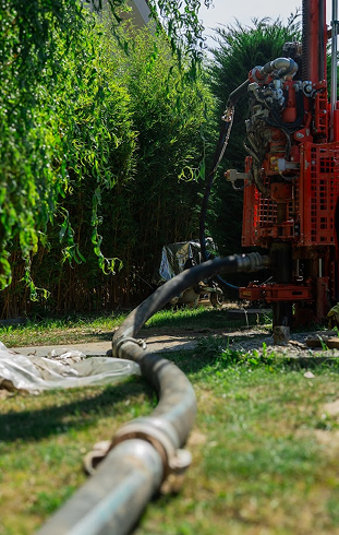 Hadice napojená na červenou vrtnou soupravu, odvádějící vyhloubený materiál z prohlubování vrtané studny na určené místo mimo pozemek.
