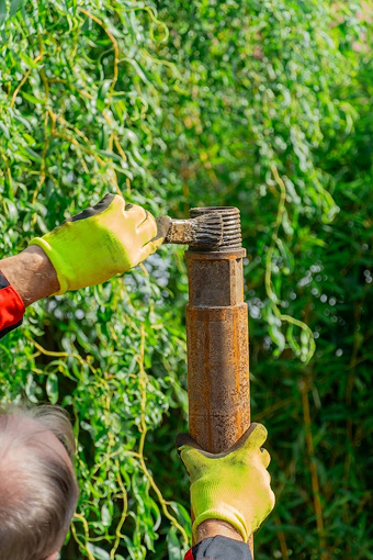 Muž v jasně žlutých ochranných rukavicích, štětcem čistí závit na horní části kovové trubky, při vrtání studny, se zeleným, rozmazaným listovím v pozadím.